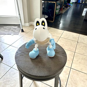 May include: A white and blue Dry Bones figurine from the Super Mario video game series. The figurine is posed on a dark wooden stool with a metal frame. The Dry Bones figure has blue gloves and shoes, and yellow eyes.