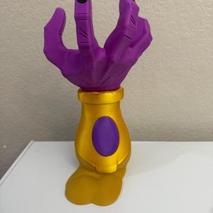 Puede incluir: Una escultura en forma de mano púrpura con uñas negras, sobre una base dorada. La base presenta un acento ovalado púrpura. Un artículo decorativo único.