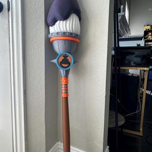 Può includere: Un bastone decorativo alto con una parte superiore viola scuro, una sezione centrale bianca e dettagli grigi e arancioni. Il bastone presenta un emblema circolare con un design a lanterna di zucca. Il manico è marrone con dettagli arancioni. Per Halloween.