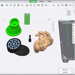 May include: A computer screen showing 3D modelling software with several 3D-rendered objects. These include a green base, a black circular object with blue lights, and a tan hand. The software interface displays tools and settings for 3D printing.