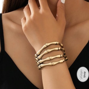May include: Gold-colored skeleton hand bracelet. The bracelet is designed to look like a hand made of bones. The bracelet is worn on the wrist. The background is a black top.