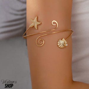 May include: Gold-coloured arm cuff with a starfish and seashell design. The cuff wraps around the arm with decorative swirls. A small pearl accents the seashell. The cuff is worn on the arm.