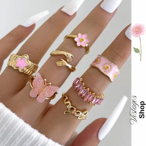 Könnte beinhalten: Eine Sammlung goldfarbener Ringe mit verschiedenen Designs, darunter ein rosa Stern, ein Schmetterling, eine Blume und ein Band mit rosa Edelsteinen. Die Ringe werden auf einer Hand mit langen, weißen Fingernägeln gezeigt. Die Ringe sind von Vistages Shop.