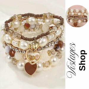 Peut inclure: Plusieurs bracelets de perles avec des accents dorés et des breloques en forme de cœur. Les bracelets présentent diverses textures, notamment des perles marron, des cristaux transparents et des perles de couleur crème. Une petite vignette montre les bracelets portés.