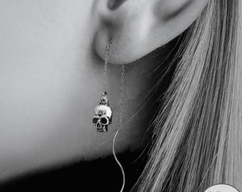 May include: A silver skull earring with a chain that dangles from the ear. The earring is worn by a person with long, straight, light-colored hair. The earring has a small skull pendant and a curved, decorative chain.