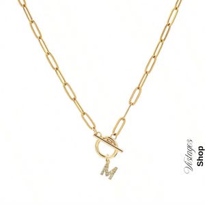 May include: Gold-coloured chain necklace with an oval link design. The necklace has a toggle clasp and a pendant in the shape of the letter "M" encrusted with small, clear crystals. The word "Vistages Shop" is printed vertically on the right side.