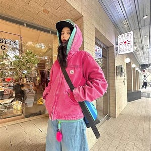 Può includere: Una persona indossa una giacca con cappuccio rosa con fodera turchese, un berretto da baseball nero e jeans azzurri. Sono visibili anche una borsa a tracolla nera e un portachiavi rosa. Lo sfondo mostra una vetrina e un'insegna con la scritta "Otter's Pocket."