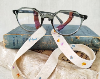 May include: Gray eyeglasses with a round frame rest on a stack of vintage books. A cream-colored ribbon with embroidered flowers and leaves is draped across the books and glasses. The books have blue covers.