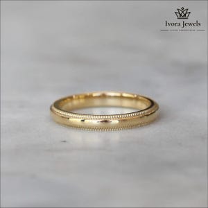 Può includere: Una fede nuziale d'oro con un bordo perlato. L'anello è posto su una superficie di marmo. La fascia ha una finitura lucida e uno stile classico ed elegante. L'immagine include il testo "Ivora Jewels".