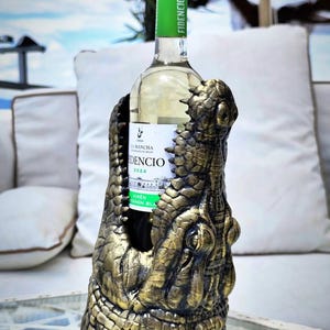 May include: A wine bottle is held in a decorative holder shaped like a crocodile's head. The holder is gold and brown, with detailed scales and texture. The wine bottle has a green label and a green top.
