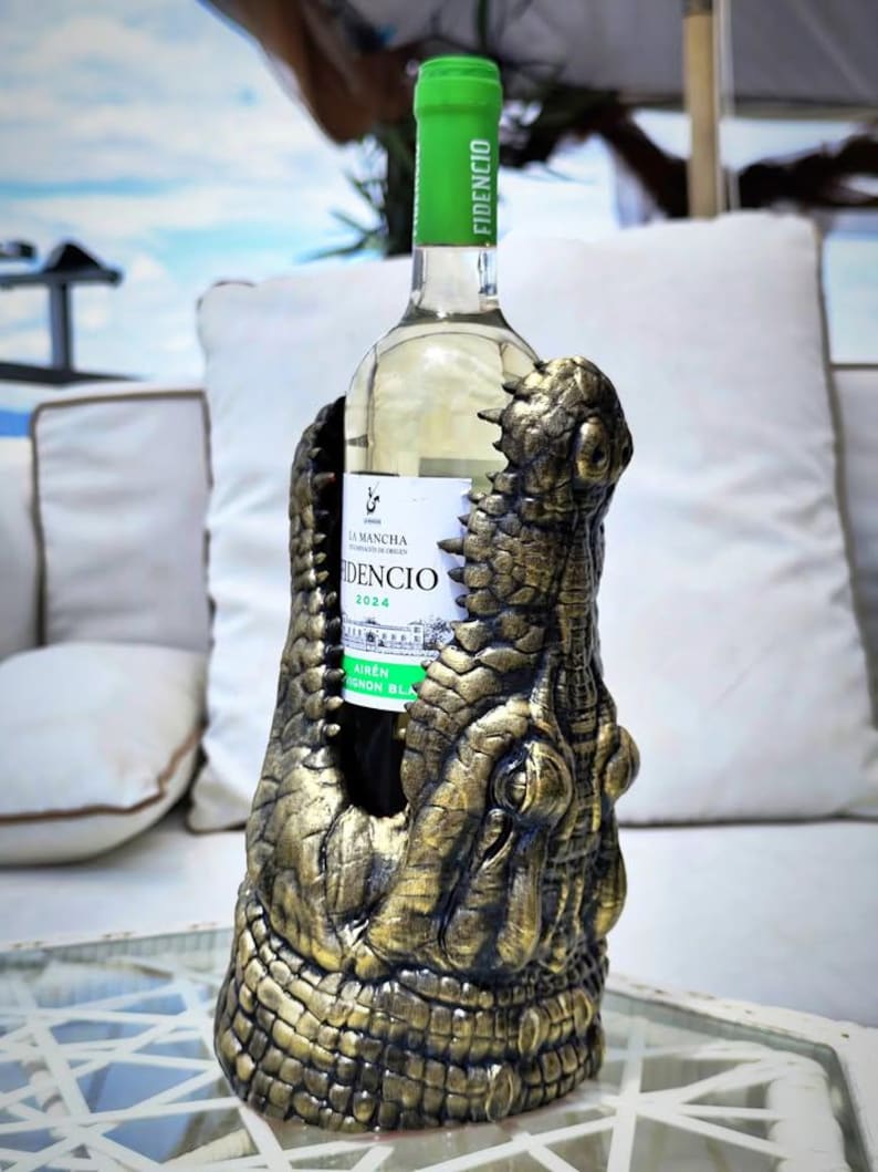 May include: A wine bottle is held in a decorative holder shaped like a crocodile's head. The holder is gold and brown, with detailed scales and texture. The wine bottle has a green label and a green top.