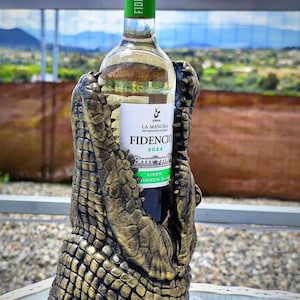 May include: A bottle of white wine in a decorative holder. The holder is shaped like a crocodile's jaws, with a textured, gold and brown finish. The wine bottle has a green top and a white label with the text "FIDENCIO 2024".