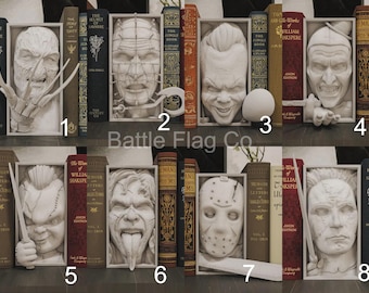 Puede incluir: Un conjunto de sujetalibros con temática de terror, con villanos icónicos del cine. Cada sujetalibros es un busto blanco esculpido de un personaje, exhibido entre libros de estilo vintage. Los sujetalibros están colocados en una estantería.