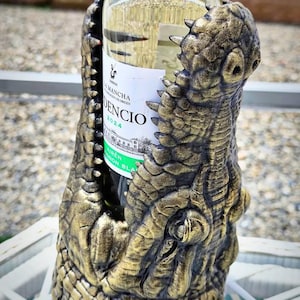 May include: A gold and bronze alligator wine bottle holder. The holder is designed to cradle a wine bottle, with the alligator's jaws open to hold the bottle. The bottle has a white label with black text.