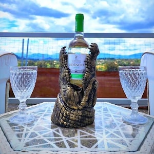 May include: A bottle of Fidencia wine in a decorative alligator wine bottle holder. Two crystal glasses sit on a white wicker table. The background features a scenic view of a blue sky and distant mountains.