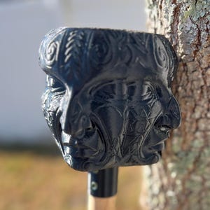 May include: A black, carved sculpture of three faces, possibly a garden tool head. The faces have detailed features and are set against a blurred natural background. The sculpture is mounted on a wooden handle.