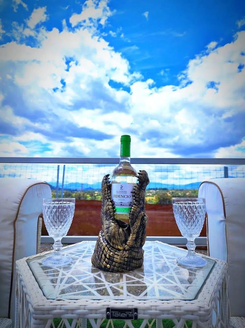 May include: A bottle of white wine in a decorative alligator wine holder, with two crystal wine glasses on a white table. The bottle has a green cap and a label that reads "FIDENCIO". The background is a bright blue sky with clouds.
