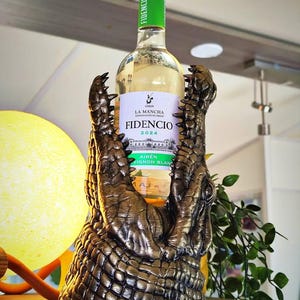 May include: A bronze-colored alligator-shaped wine bottle holder. The holder is designed to cradle a wine bottle, with the alligator's jaws open to hold the bottle. The bottle has a green cap and a white label with the text "Fidencio".