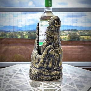 May include: A decorative wine bottle holder in the shape of a crocodile, with a gold and bronze finish. The bottle has a green cap with the word "FIDENCIO" printed on it. The bottle is on a white table.
