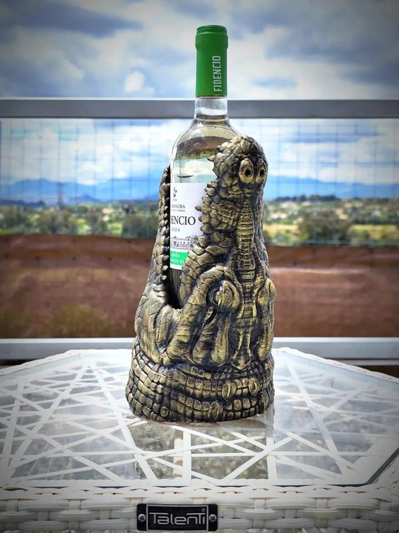 May include: A decorative wine bottle holder in the shape of a crocodile, with a gold and bronze finish. The bottle has a green cap with the word "FIDENCIO" printed on it. The bottle is on a white table.