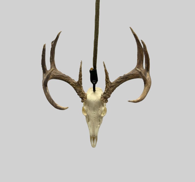 May include: A deer skull pendant with attached antlers. The skull is off-white, and the antlers are a light brown. A dark green cord is attached to the skull for hanging. The pendant is displayed against a light gray background.