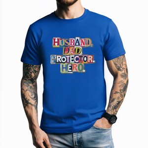 May include: A royal blue t-shirt featuring the words "HUSBAND. DAD. PROTECTOR. HERO." in a colourful, cut-out style. The text is arranged in a stacked format. The shirt is a crew neck style.
