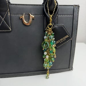 May include: A decorative bag charm featuring a cluster of green, teal, and white beads attached to a gold-tone chain and lobster clasp. The charm hangs from a black structured handbag with white stitching and a True Religion brand tag.