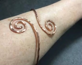 May include: A copper-colored arm cuff with a hammered texture. The cuff features a spiral design at each end, connected by a curved band. The jewelry is worn on the arm.