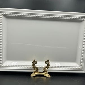 May include: A rectangular white ceramic serving tray featuring a raised beaded border along the inner edge. The tray is supported by a small, ornate, gold-colored metal display stand with curved, scroll-like details at the front base.