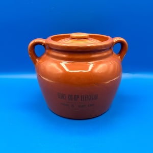 May include: A glossy orange ceramic bean pot with two side handles and a matching lid. The front features black printed text that reads: BURT CO-OP ELEVATOR, PHONE 35, BURT, IOWA. The pot has a rounded body and a smooth, reflective finish.