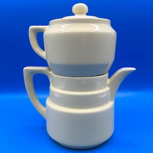 May include: A cream-colored ceramic teapot with a stacked, two-part design. The bottom section features a handle and a spout, while the top section acts as a lid with a small knob handle. The surface is smooth and glossy with a uniform finish.