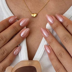 Könnte beinhalten: Eine goldene Halskette mit Herzanhänger. Die Halskette wird mit einem weißen T-Shirt getragen. Das Bild zeigt auch goldene Ringe und eine Sonnenbrille. Die Nägel sind hellrosa lackiert.