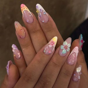 Könnte beinhalten: Eine Hand mit langen, mandelförmigen Acrylnägeln. Die Nägel sind mit floralen Designs, Glitzer und French-Tips in Pastelltönen verziert. Einige Nägel haben 3D-Blütenakzente und Strasssteine, was eine detaillierte Maniküre ergibt.