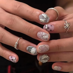 May include: Close-up of a hand with a manicure featuring various designs. The nails are painted in light purple and clear polish, with silver embellishments including flowers, stars, and a cat face. Several silver rings adorn the fingers.