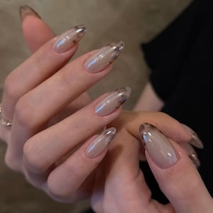 May include: Close-up of a hand with long, almond-shaped nails. The nails are painted with a nude base and a brown tortoiseshell design at the tips. The nails are glossy and reflect light.