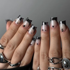 Könnte beinhalten: Nahaufnahme von Händen mit silbernen Ringen und stilvoller Nagelkunst. Die Nägel haben eine nackte Basis mit schwarzen Spitzen und Akzenten, einschließlich kleiner schwarzer Punkte und metallischer Verzierungen. Die Ringe variieren im Design.