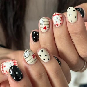 Può includere: Primo piano di una mano con manicure con vari disegni. Le unghie sono dipinte con pois bianchi e neri, un motivo a quadri rosso e bianco con un personaggio dei cartoni animati e un disegno a stella con accenti rossi.