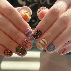 May include: A set of fingernails featuring diverse nail art designs including tiger stripes, polka dots, swirls, and abstract colour blocks. Each nail is decorated with small, metallic silver studs arranged in various patterns across the colourful polish.