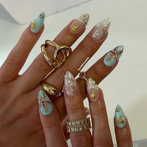Könnte beinhalten: Eine Hand, die lange, mandelförmige Nägel mit Strandmotiv zeigt. Die Nägel haben hellblaue, klare und perlmuttartige Oberflächen, verziert mit goldenen Seesternen, Muscheln und Nieten. Goldringe in verschiedenen Designs sind ebenfalls zu sehen.