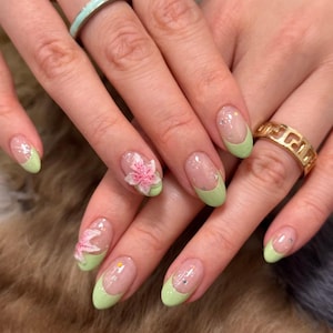 May include: Close-up of manicured nails with a light green French tip design. Some nails feature 3D pink flower accents and silver glitter. The hands also display gold and silver rings.