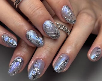Könnte beinhalten: Nahaufnahme einer Hand mit einer Maniküre mit oval geformten Nägeln. Die Nägel sind in einem hellen Lilaton mit silbernen Verzierungen lackiert. Einige Nägel haben ein silbernes Drahtdesign, andere silberne Perlen.