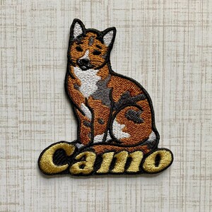 May include: An embroidered patch featuring a sitting dog with a tri-color coat of orange, white, and dark gray. The dog is positioned above the word Camo, which is stitched in gold thread with a black outline, creating a distinct graphic design.