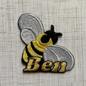 May include: An embroidered patch featuring a stylized bee with a yellow and black striped body, silver wings, and the name Ben stitched in gold thread at the bottom. The design uses black, yellow, silver, and gold embroidery thread on a fabric base.