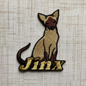 May include: An embroidered patch featuring a Siamese cat with a tan body and dark brown face, ears, and tail. The cat is sitting upright, and the word Jinx is embroidered in gold lettering with a black outline at the base of the cat figure.