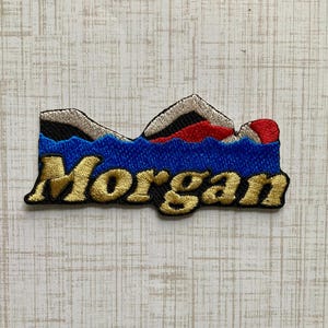May include: An embroidered patch featuring the name Morgan in gold thread against a blue background. Above the text, the design depicts stylised mountain peaks in black, white, and red colours. The patch has a distinct, irregular shape.