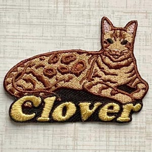 May include: An embroidered patch featuring a brown and tan spotted cat lying down. Below the cat, the word Clover is stitched in large, bold, yellow letters against a dark brown background. The design has a detailed texture with visible thread.