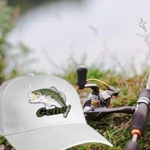 Puede incluir: Gorra de b&eacute;isbol blanca con un pez de boca grande bordado en verde y el nombre "Gene" en la parte delantera.