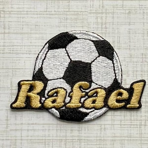 May include: A circular embroidered patch featuring a classic black and white football pattern. Overlaid across the lower portion of the ball is the name Rafael stitched in a bold, metallic gold thread with a distinct textured finish.