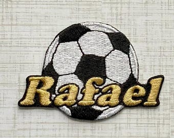 Könnte beinhalten: Ein kreisförmiger, bestickter Aufnäher mit einem klassischen schwarz-weißen Fußballmuster. Über den unteren Teil des Balls ist der Name Rafael in einem kräftigen, metallisch goldenen Garn mit einer deutlich texturierten Oberfläche gestickt.