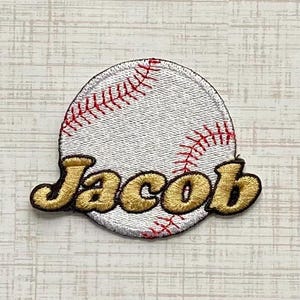 Könnte beinhalten: Ein runder, bestickter Aufnäher in Form eines weißen Baseballs mit roten Nähten. Der Name Jacob ist mittig in einer kräftigen, goldfarbenen Schrift mit dunkelbrauner Umrandung eingestickt, was dem Stoff eine strukturierte Optik verleiht.